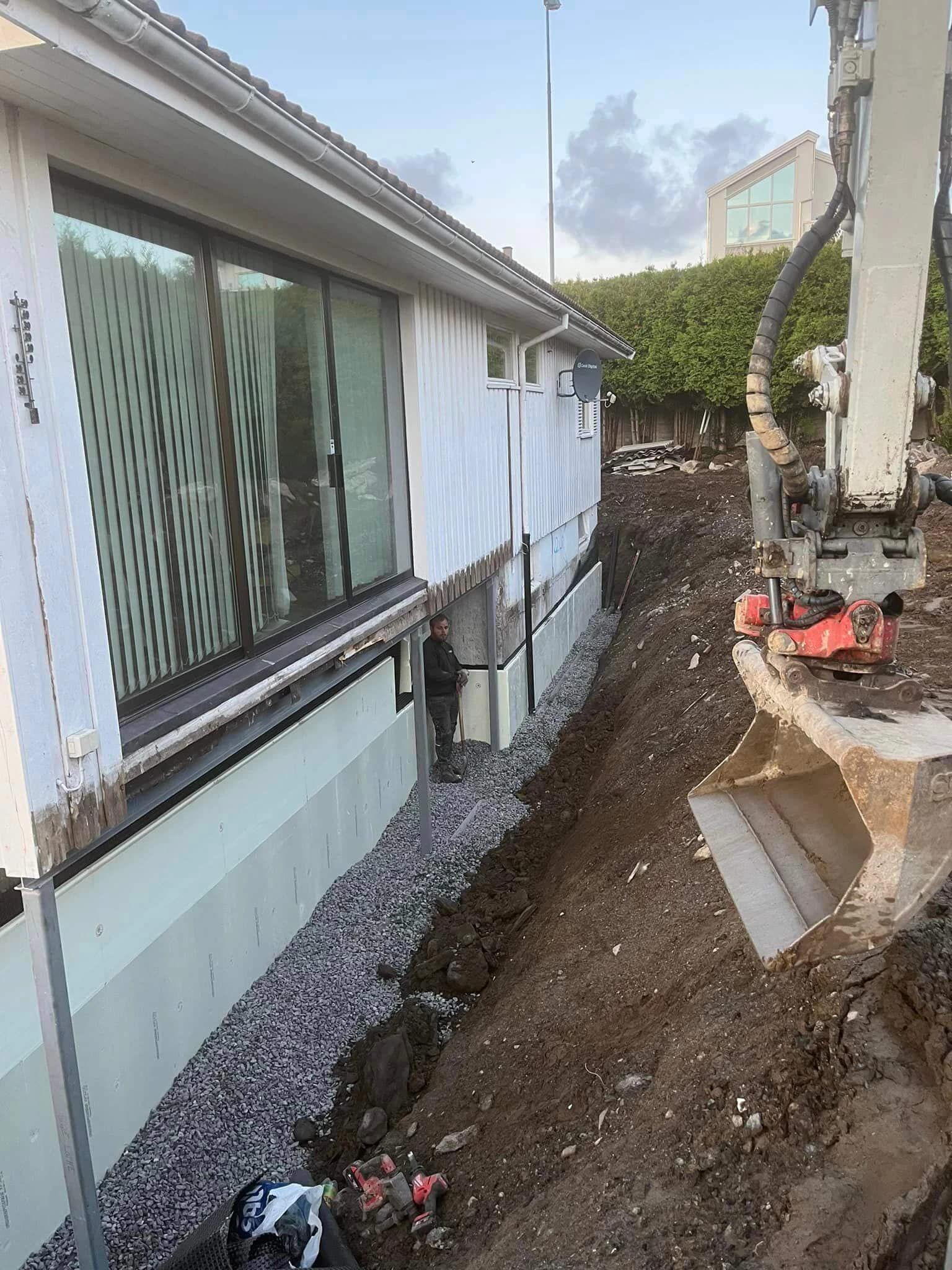 Gravemaskin og åpen dreneringsgrøft mellom en bolig og hagen.