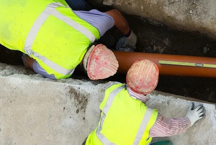 To arbeidere i synlighetsvester ved et oransje glatt rør og vater.