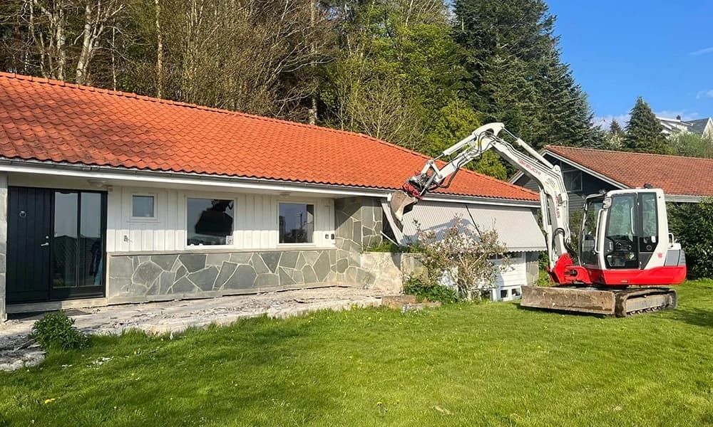 Gravemaskin ved et lavt hus med rød takstein og skadet vindusåpning.