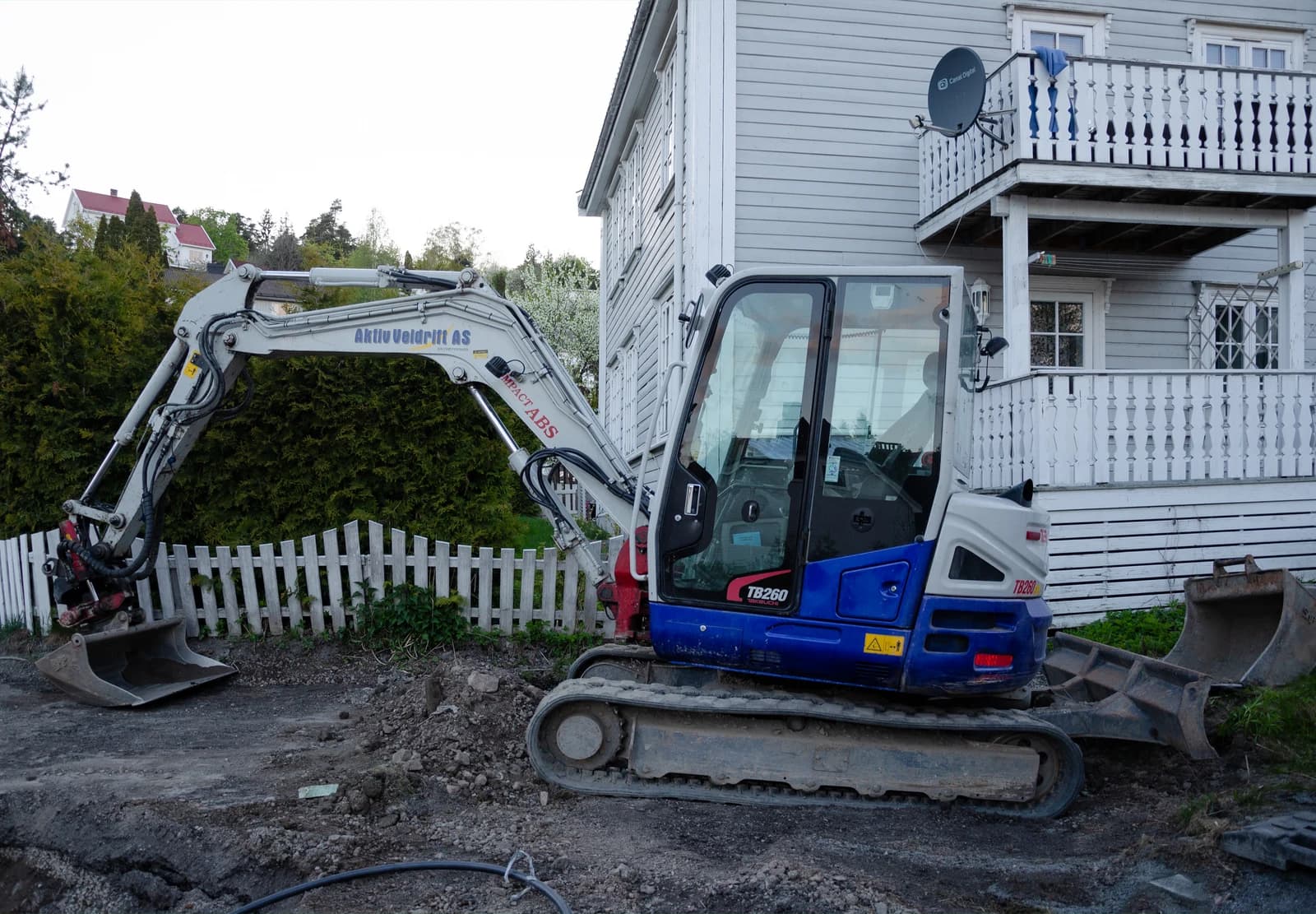Kompakt gravemaskin ved et hvitt trehus og gjerde.